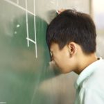 A closeup of a young student leaning his face up against a chalkboard, his eyes closed in frustration.