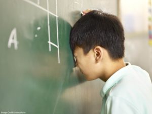 A closeup of a young student leaning his face up against a chalkboard, his eyes closed in frustration.