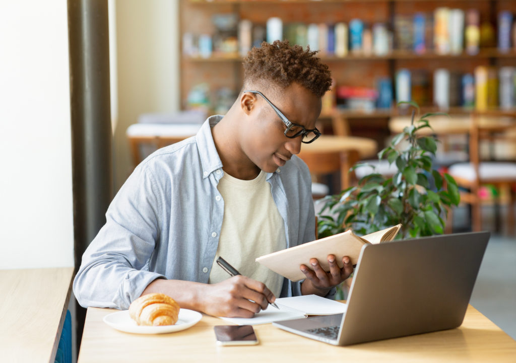 Handsome black guy in glasses learning remotely on laptop, taking notes in copybook, studying online at city cafe. Serious millennial youth working on important business project at coffee shop