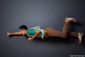 A student appears to be flying like a superhero in mid-air, but he is actually lying on his side against a dark gray background. He extends one arm forward in a classic "superhero flight" pose, while his legs are bent, creating the illusion of movement. He wears brown pants, a leather belt, and sneakers. The creative perspective and lighting make it look as if he is defying gravity.