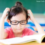 A young student looks at a book open on her desk and scratches her head in confusion