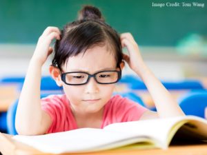 A young student looks at a book open on her desk and scratches her head in confusion