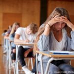 Students sitting in a gym at desks, presumably taking exams. The student in front shields her eyes with her hands, looking stressed and depressed.