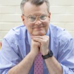 Photograph of the author, wearing a blue shirt, pink tie, and glasses, smiling at the camera