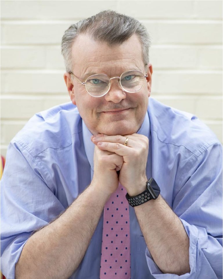 Photograph of the author, wearing a blue shirt, pink tie, and glasses, smiling at the camera