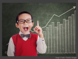 Child wearing a bow tie and a happy expression standing in front of a chalkboard with a bar graph showing steady increases