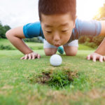 Little boy blowing golf ball into hole.