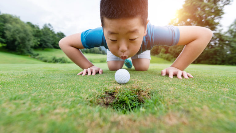 Little boy blowing golf ball into hole.