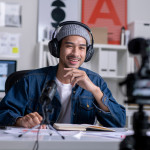 A smiling young man wearing a jeans jacket, wool cap, and headphones sits at a desk and talks to a camera in front of him.
