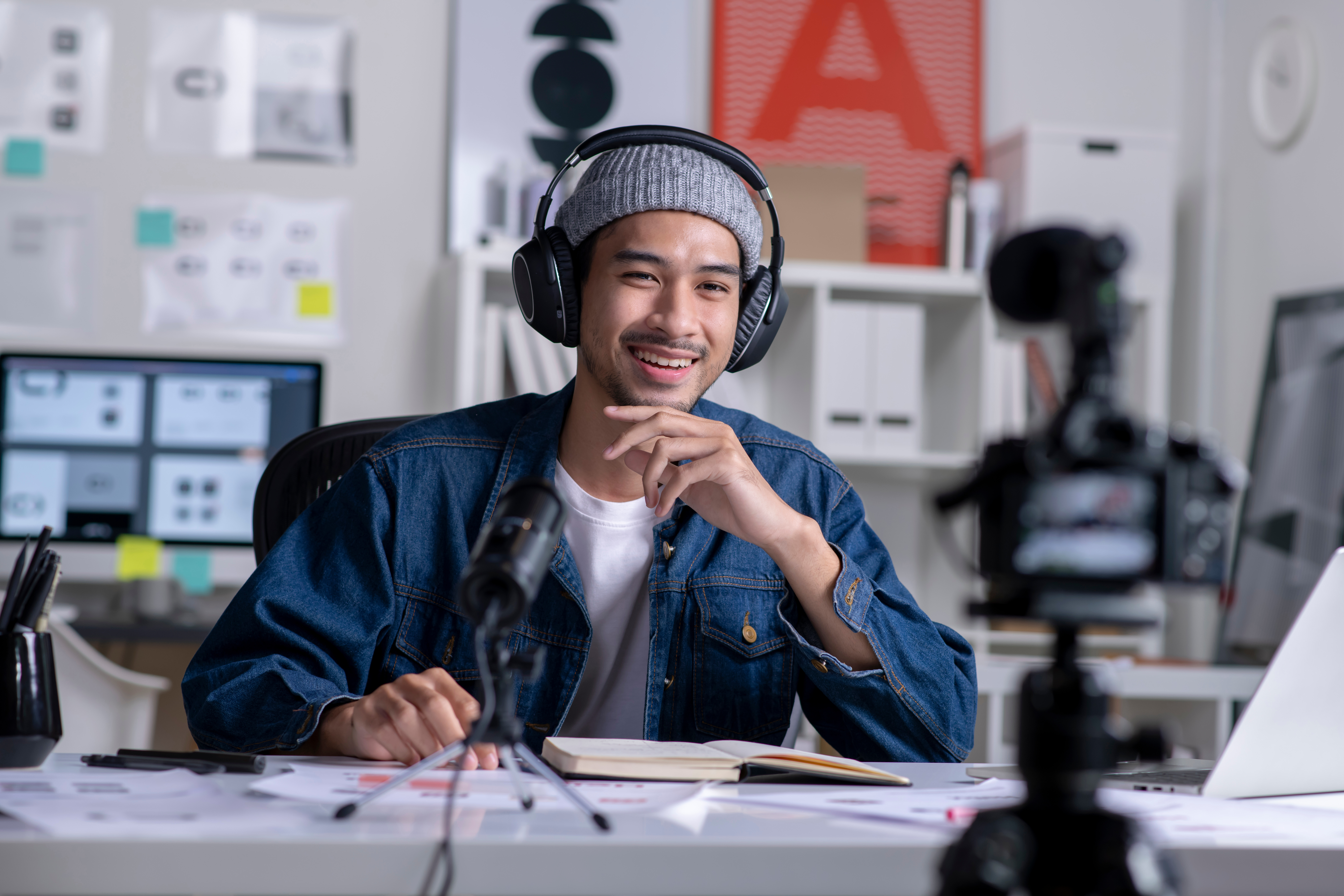A smiling young man wearing a jeans jacket, wool cap, and headphones sits at a desk and talks to a camera in front of him.