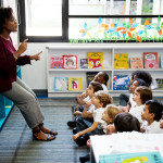 Kindergarten students sitting on the floor, listening to the teacher at the chalkboard