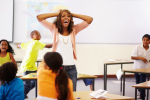 Exasperated teacher standing in the middle of a chaotic classroom, holding her hands on her head and shouting