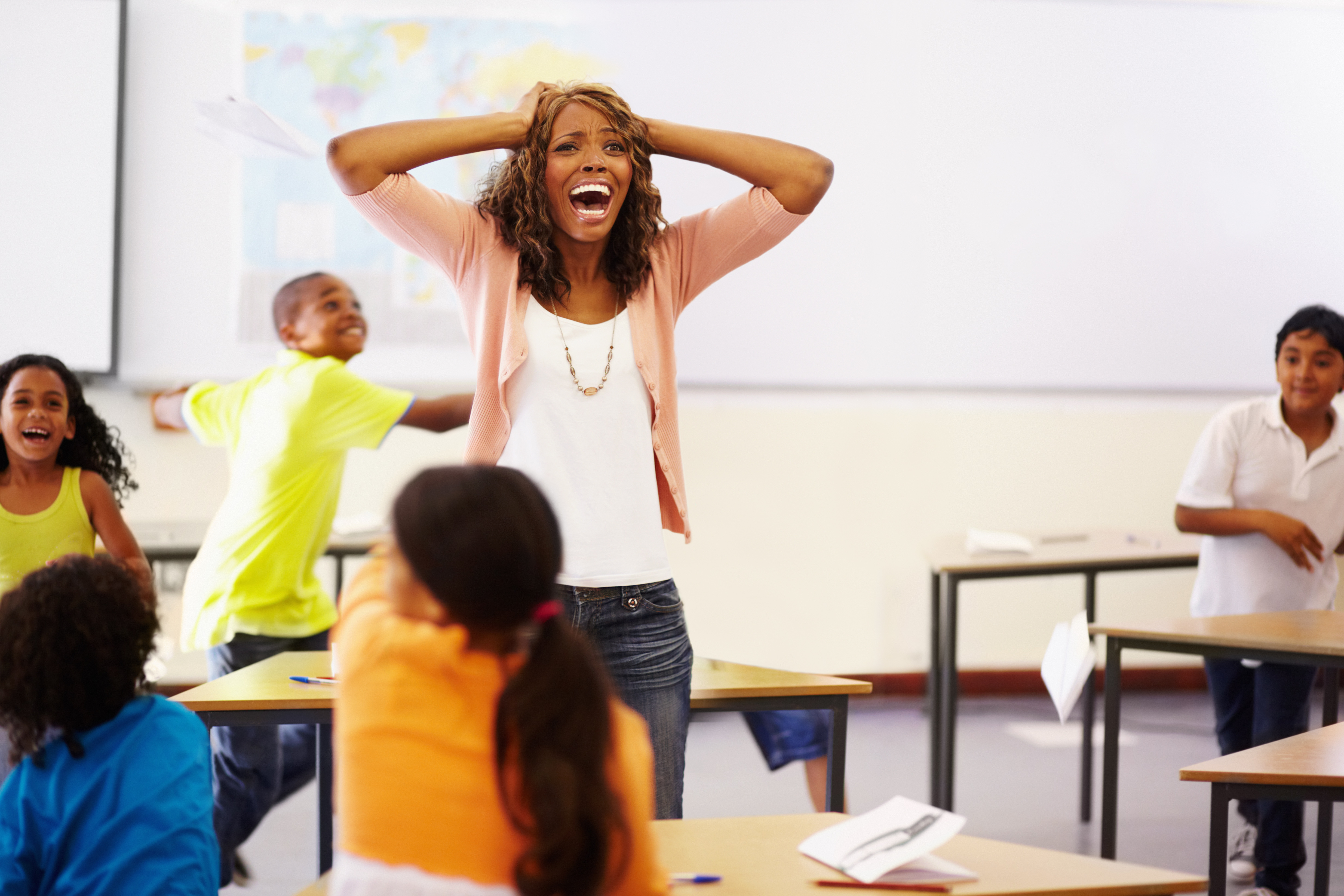 Exasperated teacher standing in the middle of a chaotic classroom, holding her hands on her head and shouting