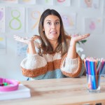 Young teacher wearing sweater and glasses sitting on desk at kindergarten clueless and confused expression with arms and hands raised.