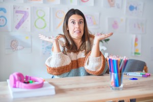 Young teacher wearing sweater and glasses sitting on desk at kindergarten clueless and confused expression with arms and hands raised.