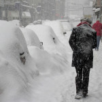 A winter scene: cars covered in a foot of swon, and two pedestrians walking away from the camera, shoulders hunched agains the cold snow