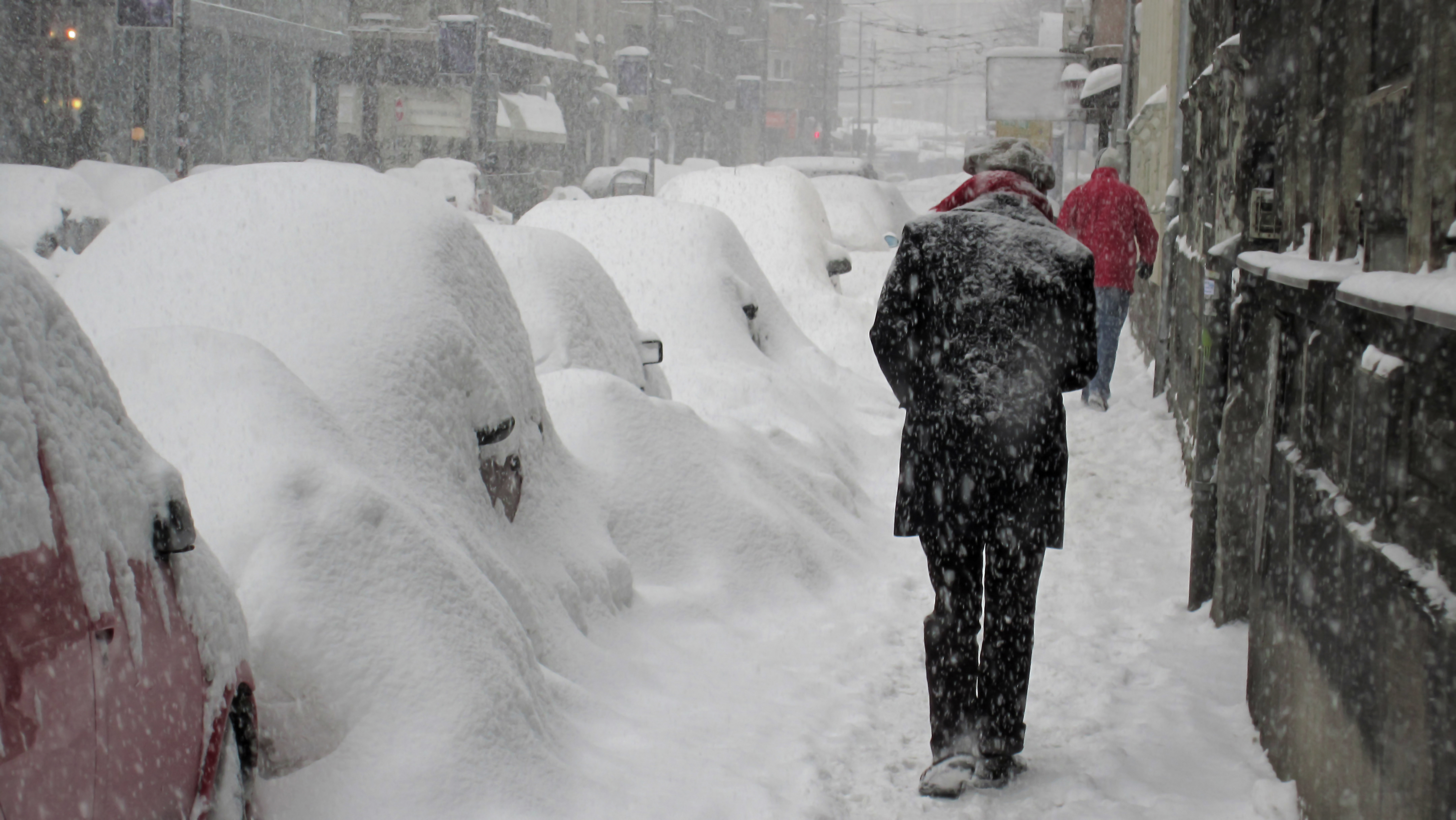 A winter scene: cars covered in a foot of swon, and two pedestrians walking away from the camera, shoulders hunched agains the cold snow