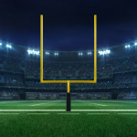 A bright yellow American football goalpost, above a bright green field and against dark stadium