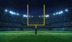 A bright yellow American football goalpost, above a bright green field and against dark stadium