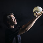An actor, presumably playing Hamlet, holding up a skull while standing against an all-black background