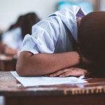 Close up of student with head down on a wooden desk, hair covering his or her face. Other students are working out of focus in the background.