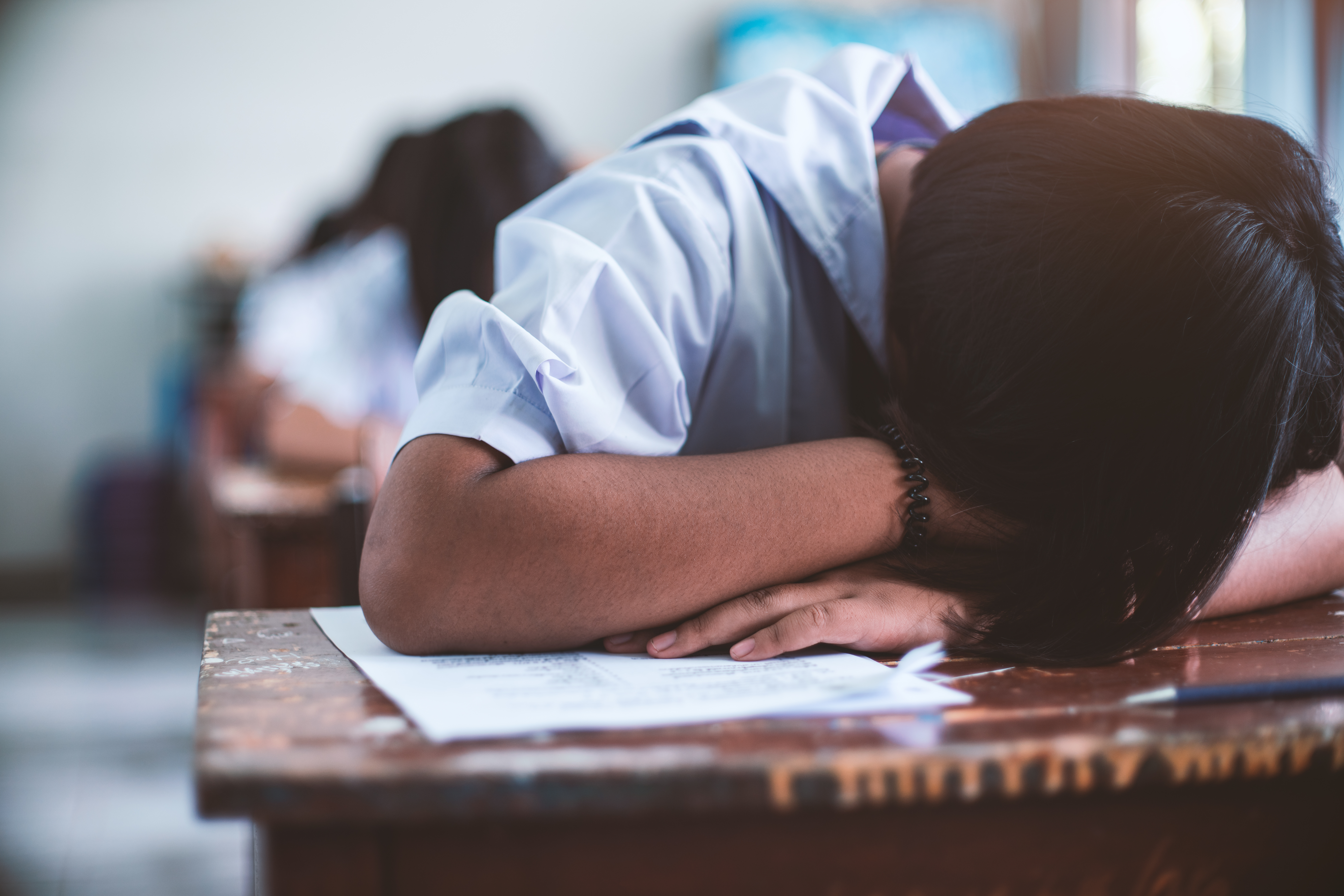 Close up of student with head down on a wooden desk, hair covering his or her face. Other students are working out of focus in the background.