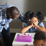 A young student sits at a desk with her hands covering her eyes; a sympathetic teacher stands next to her with his hand on her shoulder