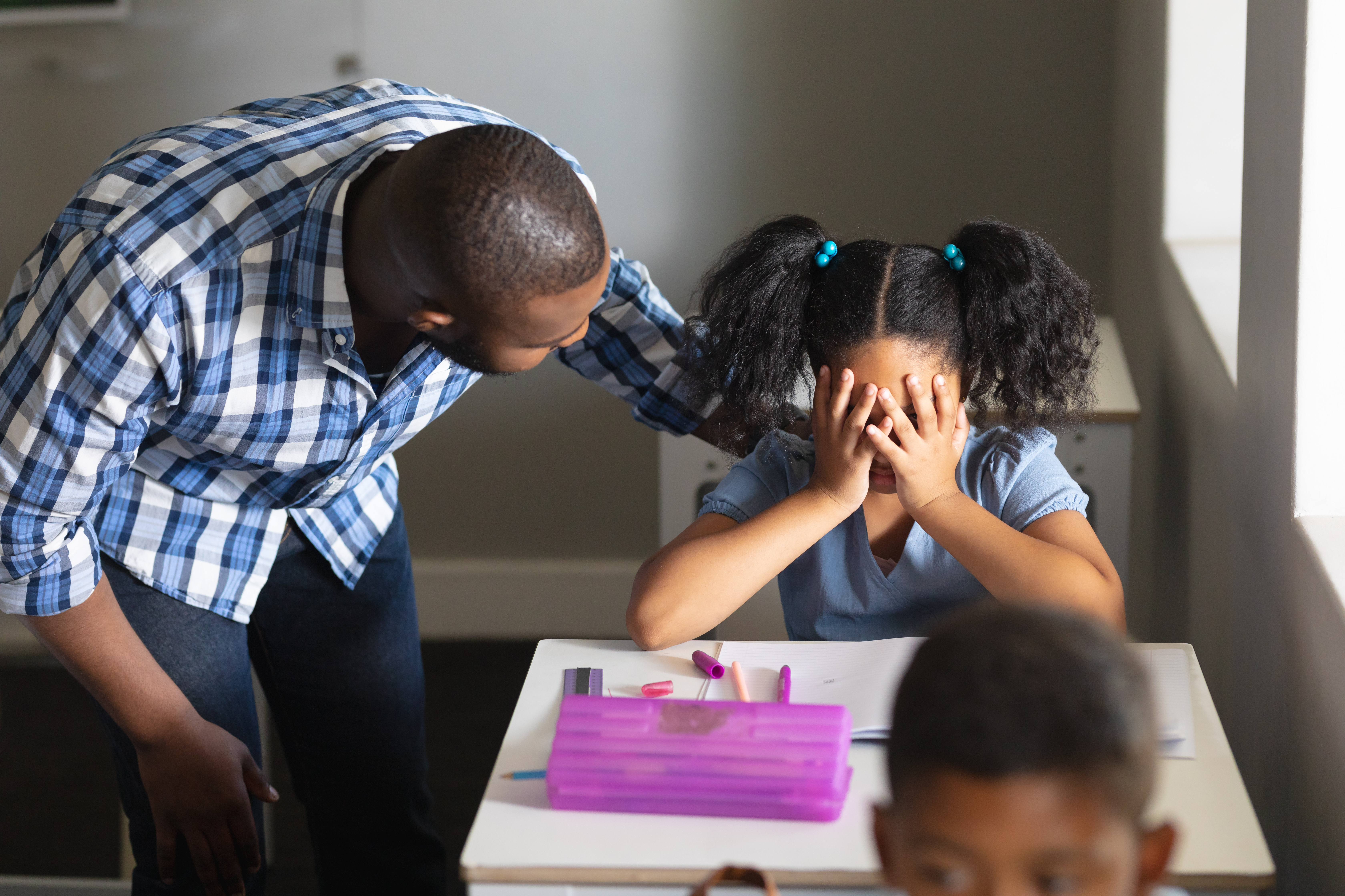 A young student sits at a desk with her hands covering her eyes; a sympathetic teacher stands next to her with his hand on her shoulder