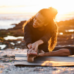 A woman doing yoga on the beach at sunset; rocks and the ocean visible in the background
