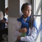 A young student with long dark hair stands pensively by a school hallway window, clutching a green notebook and wearing a blue scarf over her white uniform shirt. Her expression appears troubled or thoughtful as she gazes outside. In the background, other uniformed students interact in the corridor. The image captures a moment of isolation or contemplation within the busy school environment, suggesting themes of teenage emotional challenges in educational settings.