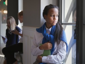 A young student with long dark hair stands pensively by a school hallway window, clutching a green notebook and wearing a blue scarf over her white uniform shirt. Her expression appears troubled or thoughtful as she gazes outside. In the background, other uniformed students interact in the corridor. The image captures a moment of isolation or contemplation within the busy school environment, suggesting themes of teenage emotional challenges in educational settings.