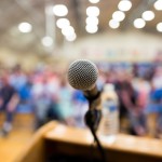 A close-up of a microphone on a podium, with the silver mesh head in sharp focus against a blurred background. Behind the microphone is a large, out-of-focus audience in what appears to be an auditorium or gymnasium with ceiling lights visible above. The wooden podium is partially visible at the bottom of the frame. The image captures the perspective of a speaker addressing a crowd, suggesting a public speaking event, conference, or school assembly.