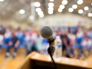 A close-up of a microphone on a podium, with the silver mesh head in sharp focus against a blurred background. Behind the microphone is a large, out-of-focus audience in what appears to be an auditorium or gymnasium with ceiling lights visible above. The wooden podium is partially visible at the bottom of the frame. The image captures the perspective of a speaker addressing a crowd, suggesting a public speaking event, conference, or school assembly.