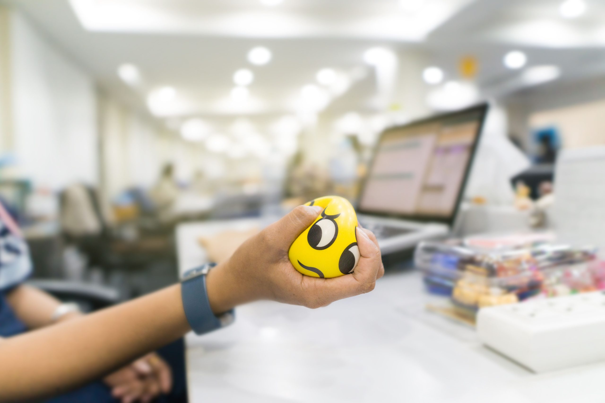 A hand squeezing a yellow squishy ball with a cartoon face on it; a computer screen and classroom in the background.